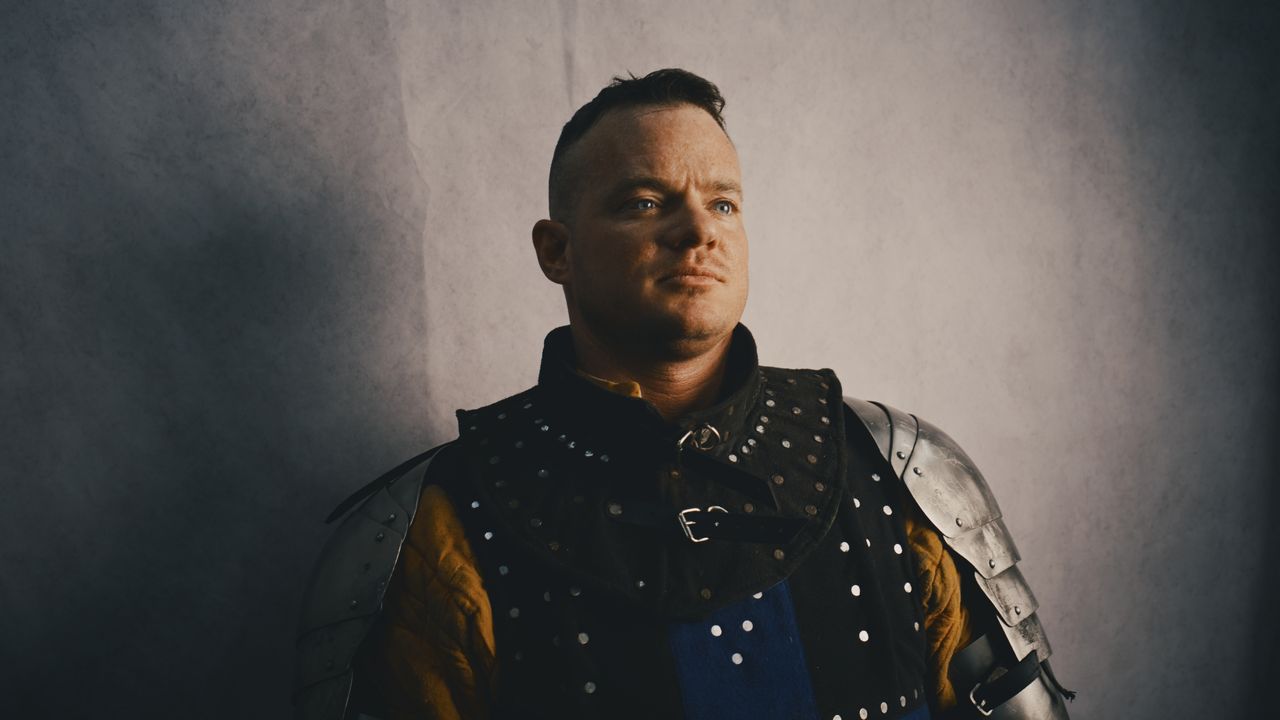 A man in a studded black gambeson and steel pauldrons looks past the camera, lit from one side against a painted grey backdrop.