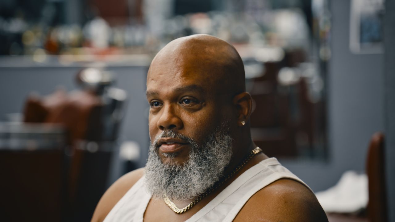 A bald man with a grey beard and gold chain speaks during an interview in a barbershop.