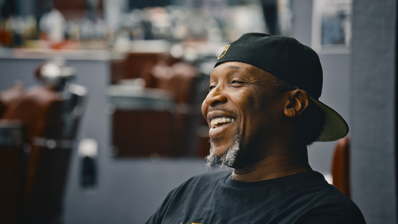 A man in a backwards black cap laughs in profile, barber chairs blurred behind him.