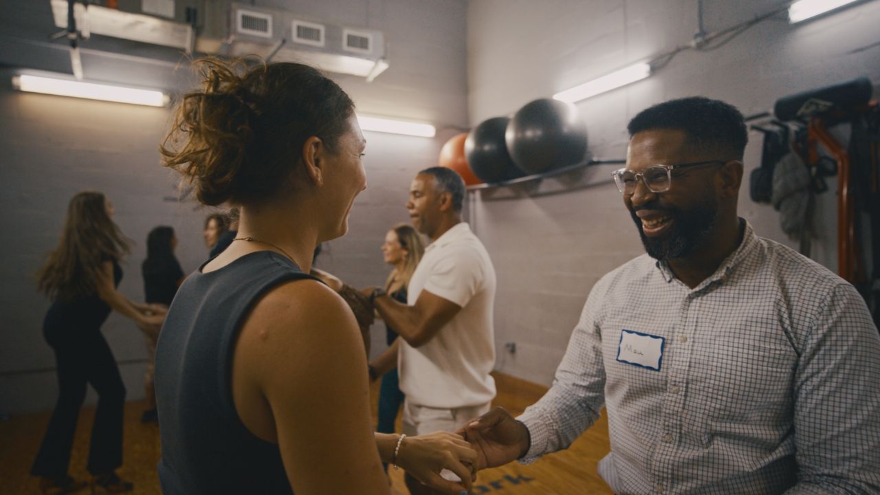 Two people laugh as they partner up in a dance class, other couples moving behind them.