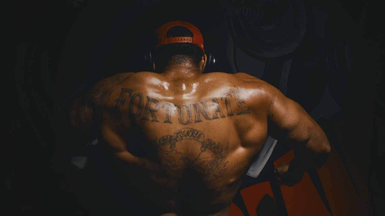 A lifter seen from behind in a dark gym, sweat on his shoulders, the word Fortunate tattooed across his back.