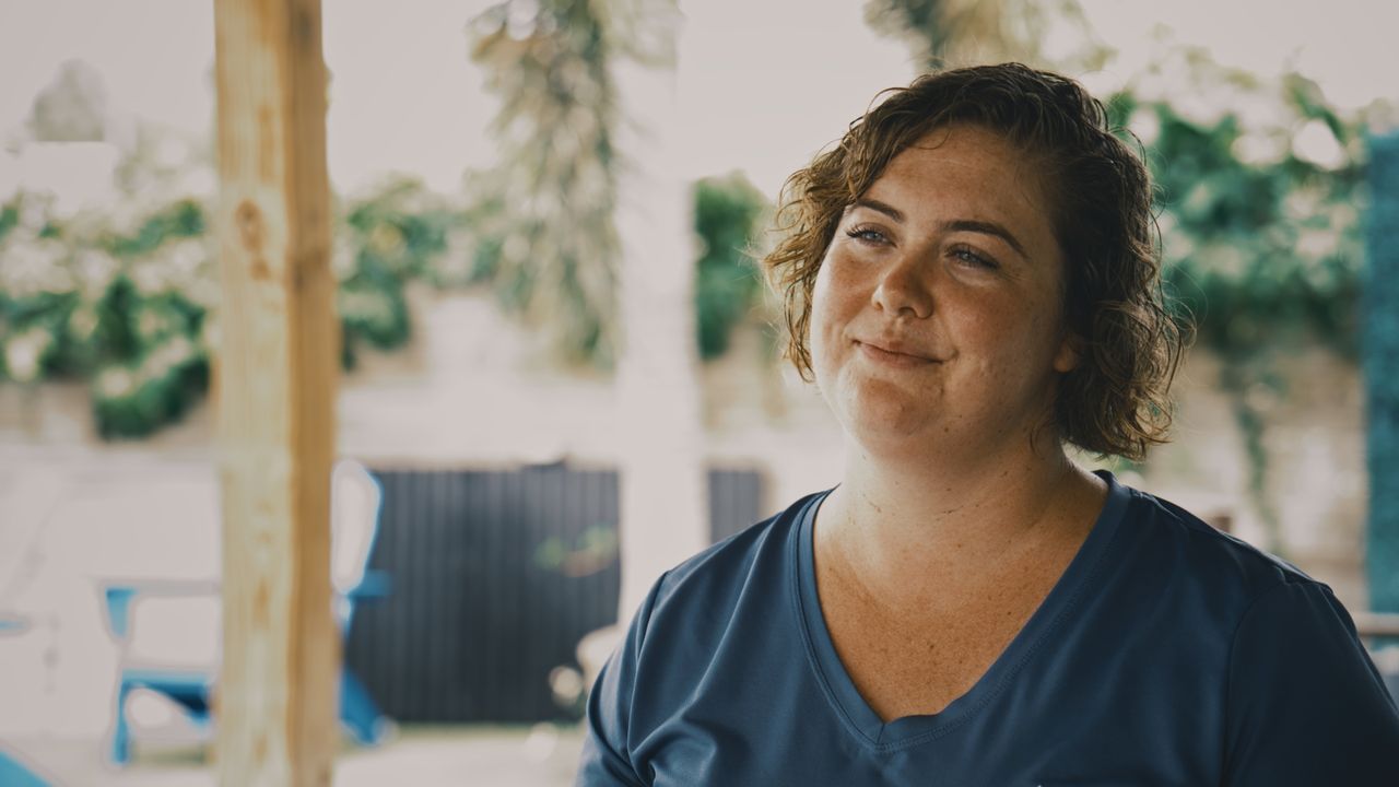 A woman with curly hair and a blue shirt smiles off camera on a covered patio.