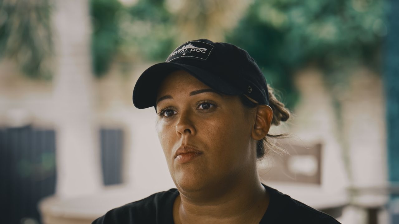 Close-up of a woman in a black Total Dog Tampa cap, listening, greenery blurred behind her.